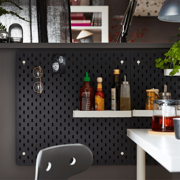 A black IKEA SKÅDIS pegboard and add-on white shelves and pegs, used to hang accessories and study and kitchen supplies. 