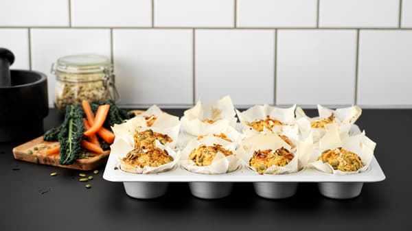 A black kitchen worktop with a VARDAGEN metal muffin tin filled with twelve muffins made with carrots and pumpkin seeds.