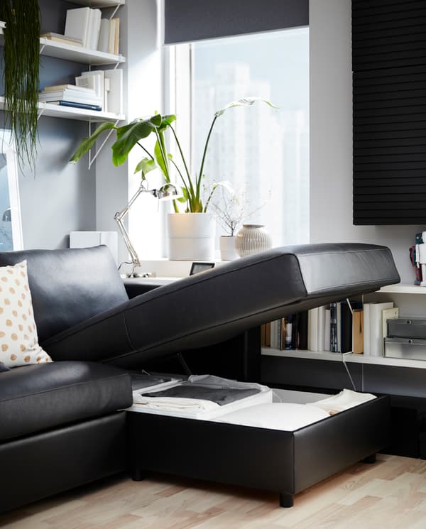 A black leather sofa-bed with a chaise longue seat that’s open, bedding stored underneath, white shelves and a green plant.
