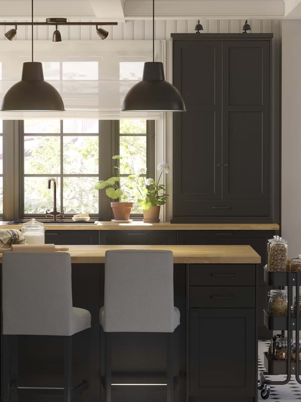 A black LERHYTTAN kitchen with two black lamp shades hanging above a kitchen island and a RASKOG cart next to it.