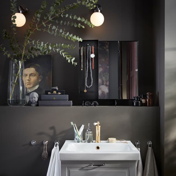 A black mirror, a vase with eucalyptus, two round wall-mounted lamps and a white wash-basin.