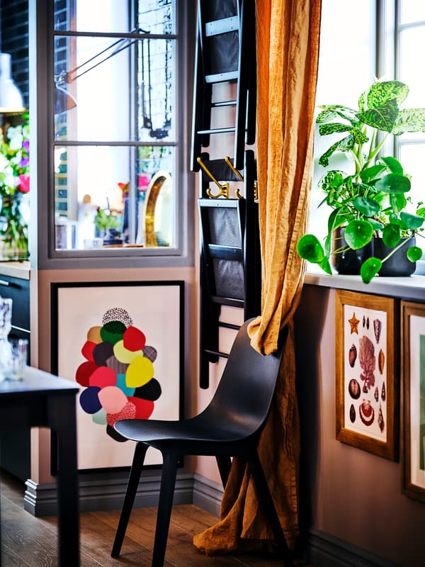 A black ODGER chair, surrounded by framed art on the walls, a folded TERJE chair and potted plants on the windowsill above.