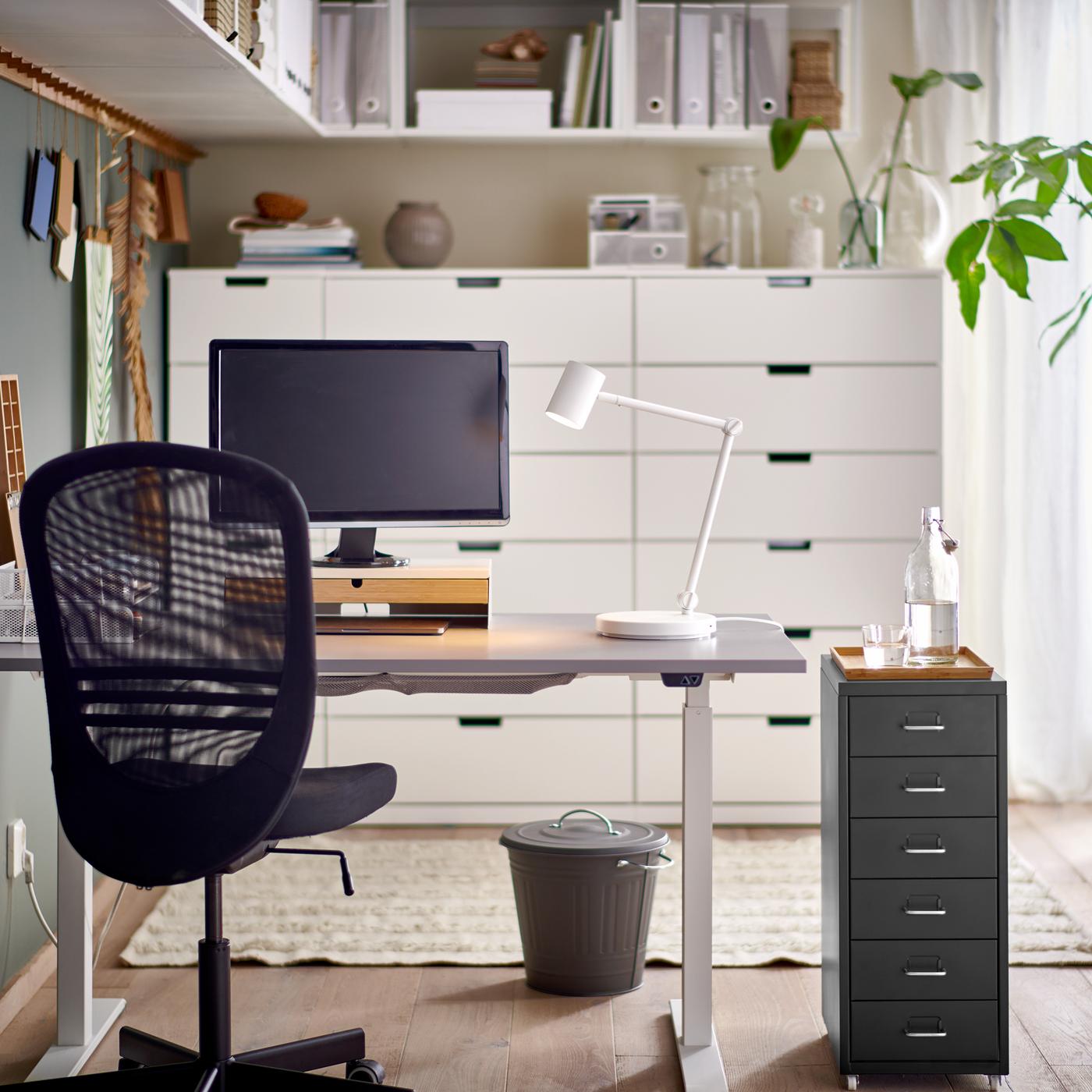 A black office chair and a HELMER drawer unit by a grey/white RODULF sit/stand desk with a work lamp and computer screen.