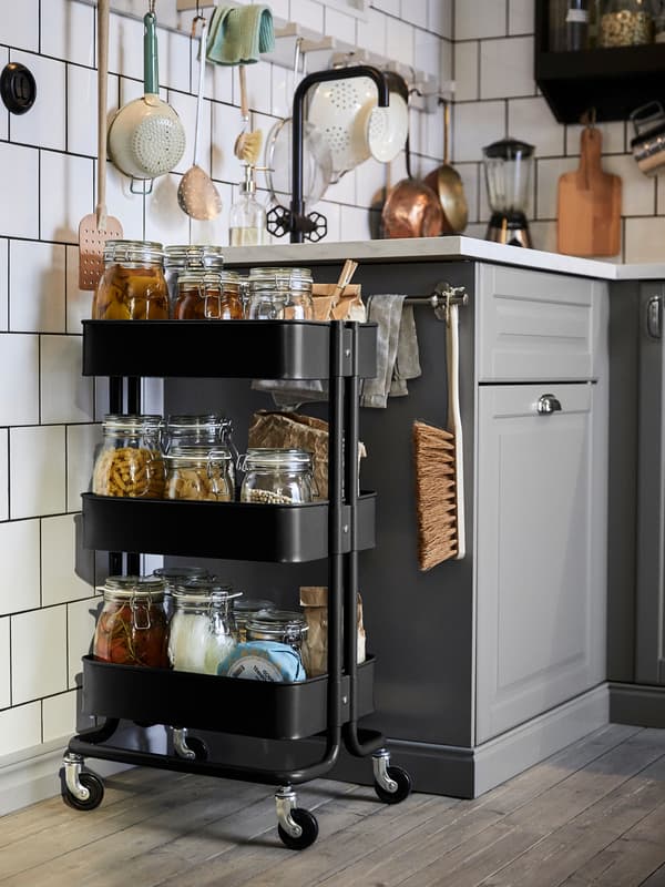 A black, RÅSKOG trolley, with glass jars of various foods on its shelves, stands next to a sink in a white-tiled kitchen.