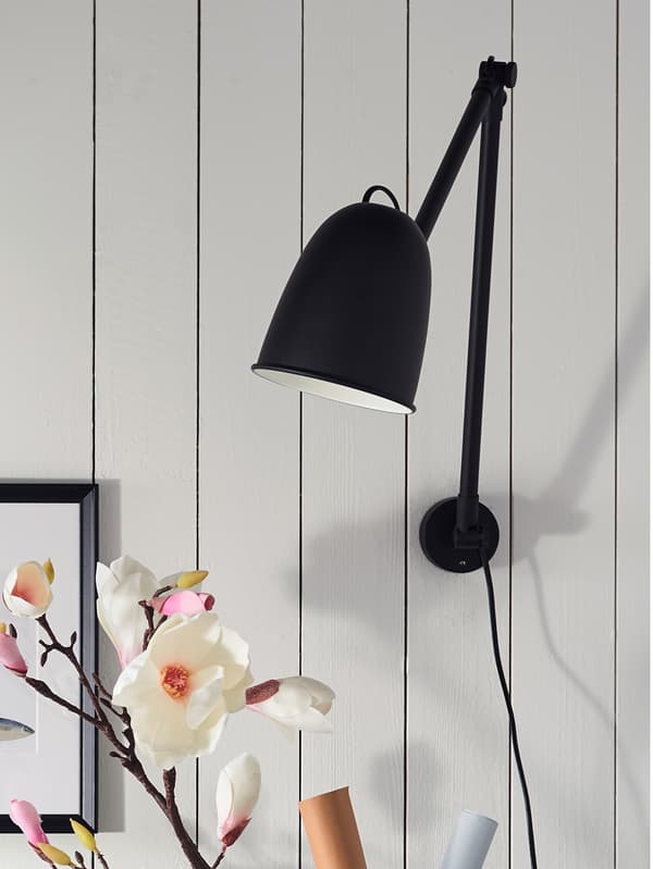 A black SKURUP work/wall lamp is attached to a wall made of wooden planks that have been painted white.