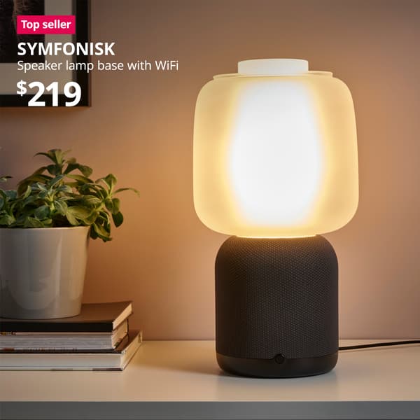 A black SYMFONISK smart speaker lamp base with Wifi and white glass shade lit and placed on the shelf. A red "Top seller" label is on the top left of image, with white text below that reads: SYMFONISK, Speaker lamp base with WiFi. Price at $219.