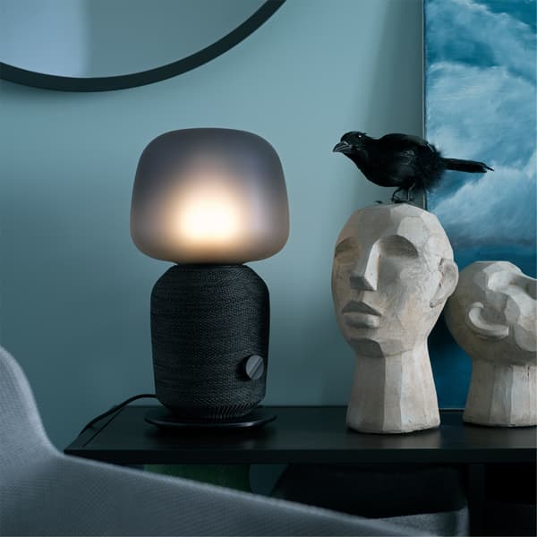A black SYMFONISK table lamp is standing on a black table next to decorative items in a blue bedroom.
