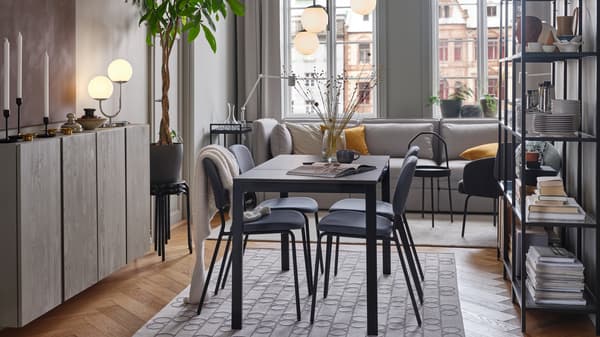 A black table and four grey chairs stand on a geometric-patterned rug. Above hangs a 3-armed chrome-plated pendant lamp.