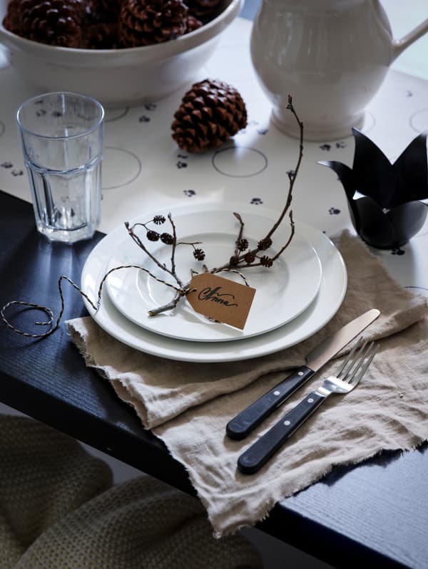 A black table is set with light-coloured dinnerware and natural accessories, including pinecones and sticks.