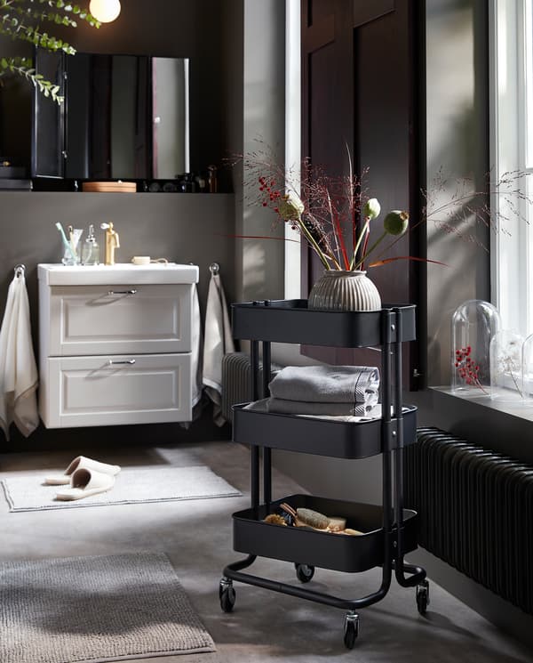 A black trolley on castors with a vase and towels on the shelves. Behind is a white wash-stand/wash-basin.
