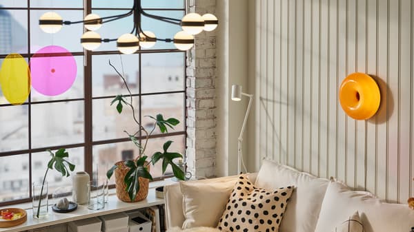 A black/white STORSLINGA chandelier in a room with a white sofa, a white floor/reading lamp and a pot plant in the window.