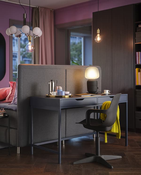 A blue ALEX desk, a lit table lamp and a black swivel chair with a yellow throw are standing at the back of a bed headboard.
