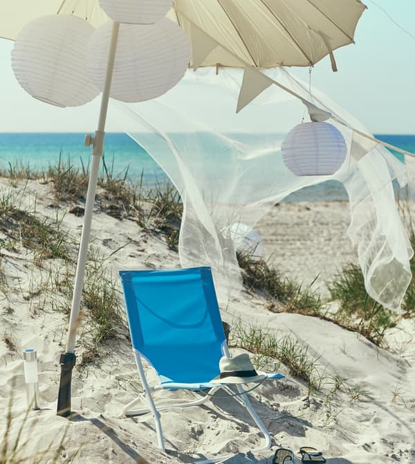 A blue beach chair sits in the sand under a large white umbrella and solar-powered pendant lamps.