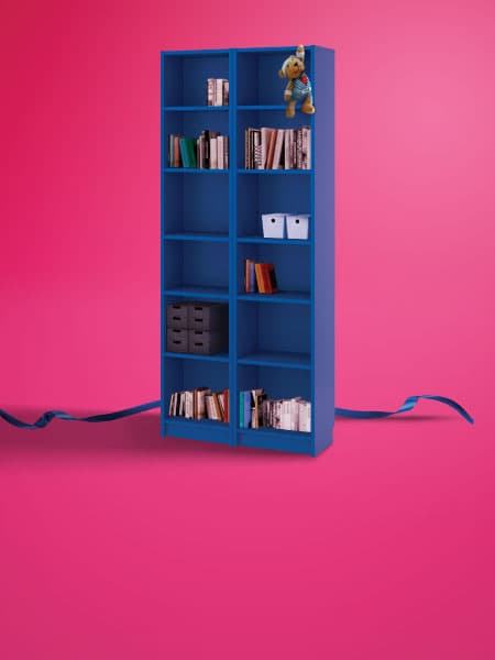 A blue BILLY bookcase with a FABLER BJÖRN soft toy bear hanging on the top right corner of the bookcase on a pink background.
