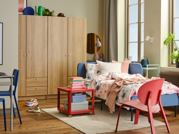 A blue BLÅKULLEN bed with corner headboard stands next to a TINGBY side table on castors near a red FRÖSET easy chair.
