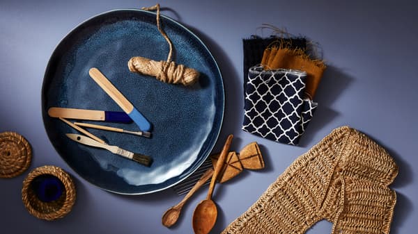 A blue ERTAPPAD dish holding a roll of string and paint brushes, beside fabric samples, wooden spoons and woven objects.