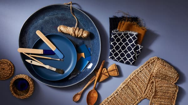 A blue ERTAPPAD dish holding a roll of string and paint brushes, beside fabric samples, wooden spoons and woven objects.