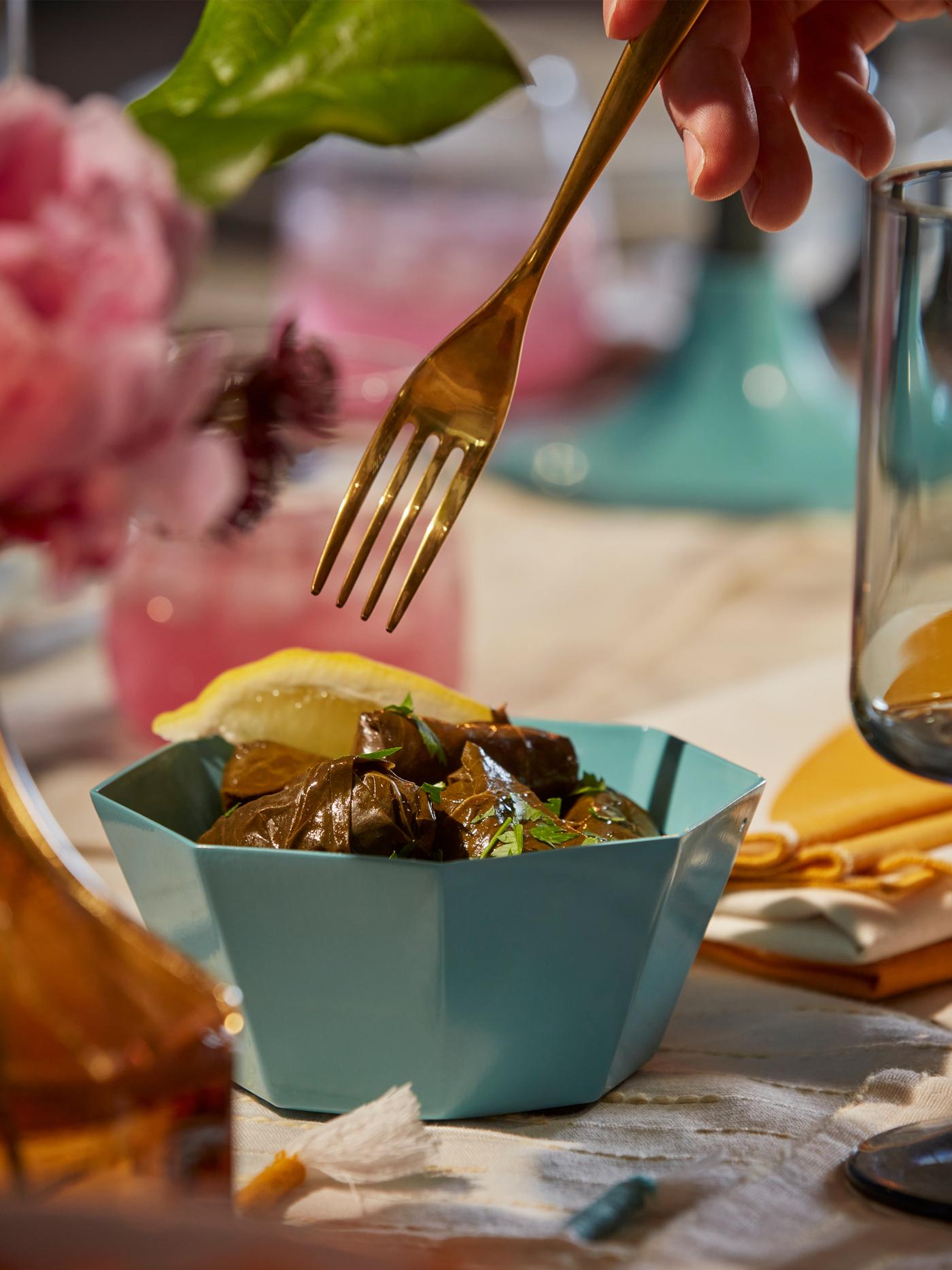 A blue GOKVÄLLÅ serving bowl with food in it on a set table. Above the bowl, a hand is holding a fork.