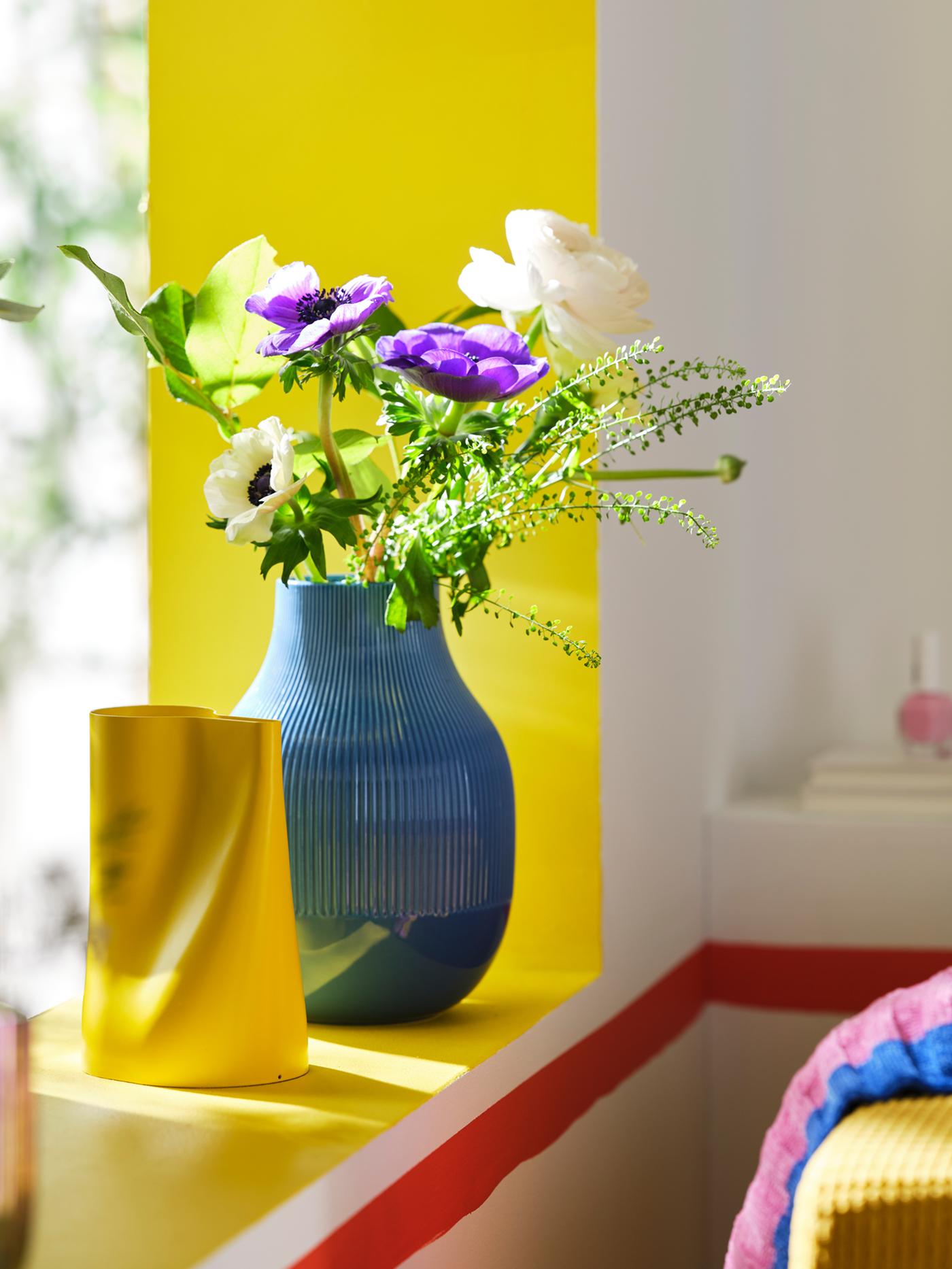 A blue GRADVIS vase with fresh flowers in it and a bright yellow CHILIFRUKT vase, both placed on a yellow windowsill.