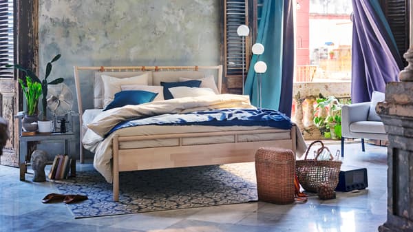 A blue-grey bedroom with a wooden double bed and turquoise and purple block-out curtains blowing in the wind.