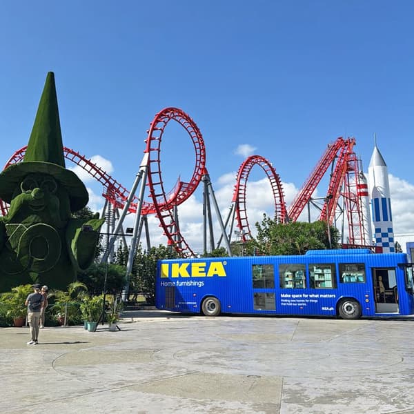 A blue IKEA bus parked in front of a large green topiary figure, with red roller coaster tracks and a rocket structure in the background.