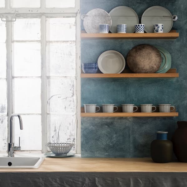 A blue kitchen space with a window and sink and open wood shelves holding dishes.