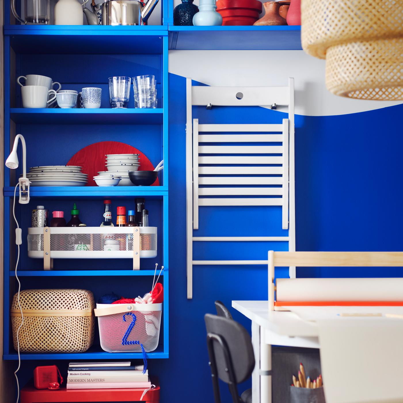 A blue PLATSA open shelving unit stores several baskets and a white FRÖSVI folding chair is hanging on hooks next to it.