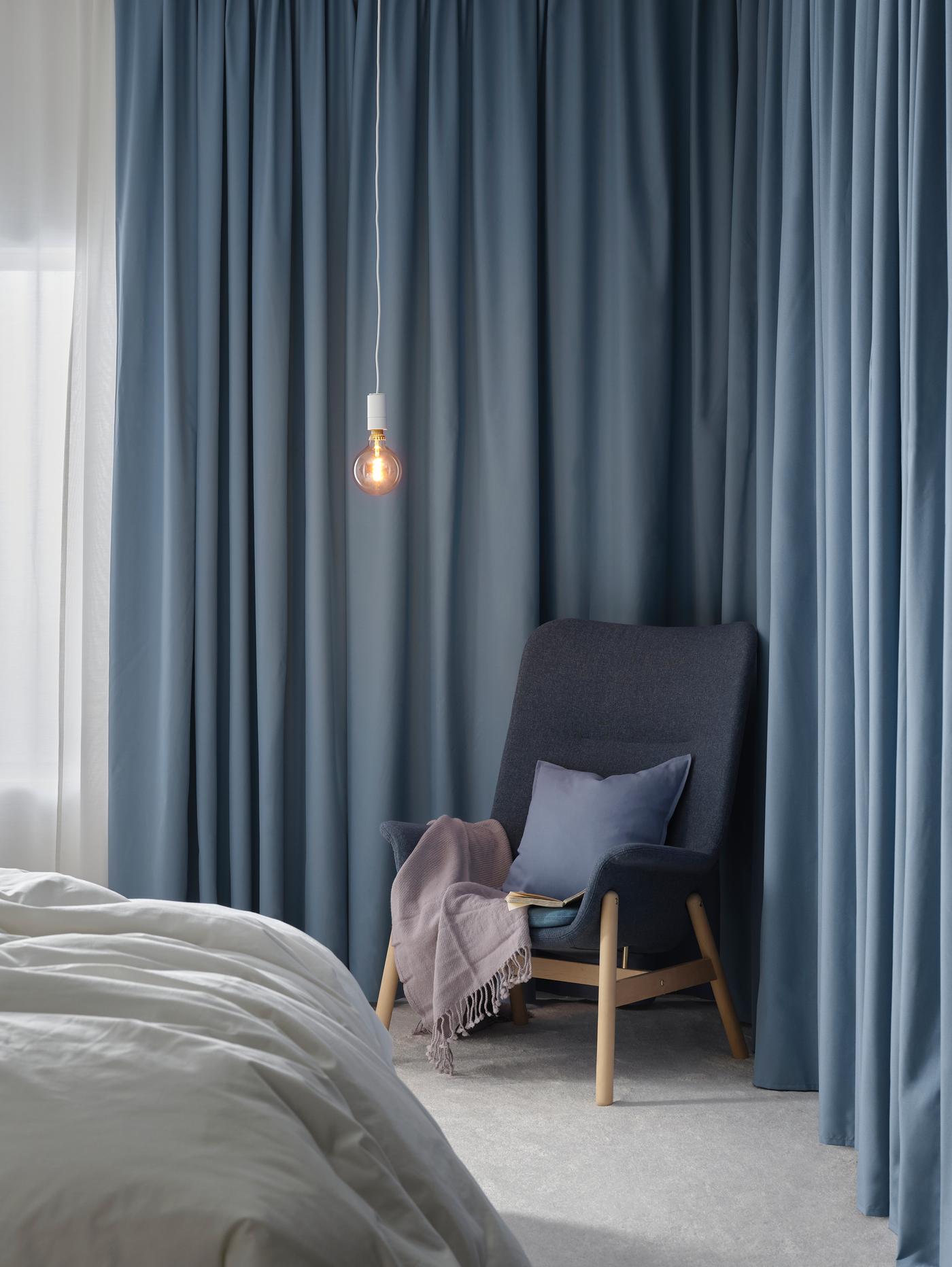 A blue, Scandinavian-styled VEDBO high-back armchair with birch legs holds a throw and cushion in front of blue curtains.