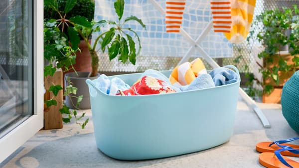 A blue TORKIS flexi laundry basket filled with laundry, a drying rack and a pouffe on a sunny balcony decorated with plants.