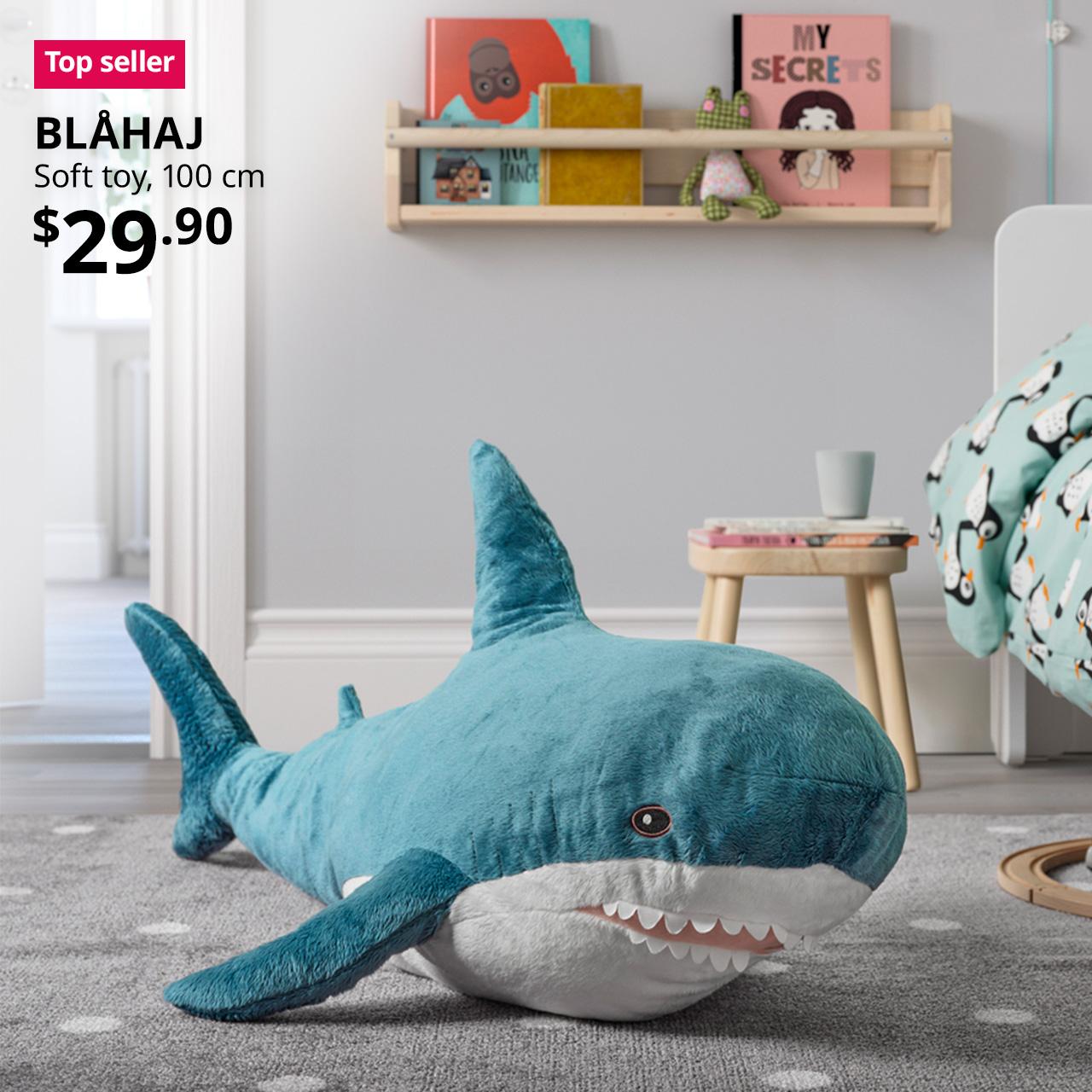 A blue/white BLÅHAJ shark soft toy on a grey with white polka dot rug in a children's bedroom. A redish pink tag with white text that reads "Top seller", followed by a black text overlay image that reads "BLÅHAJ, Soft toy, 100 cm, $29.90". 