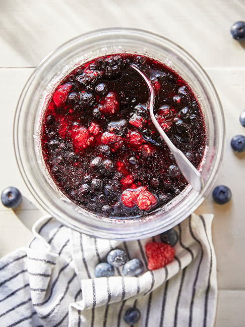 A blueberry and a strawberry rest in a creamy mixture inside a glass container, with a spoon nearby highlighting fresh ingredients.