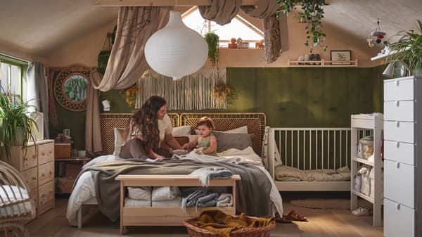 A boho feel parent and baby shared bedroom with mum and baby sitting on a double bed with a white GULLIVER cot beside it.