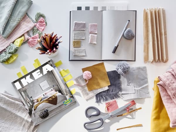 A bookmarked IKEA catalogue, an open notebook, a cup of coloured pencils and fabric samples in various colours.