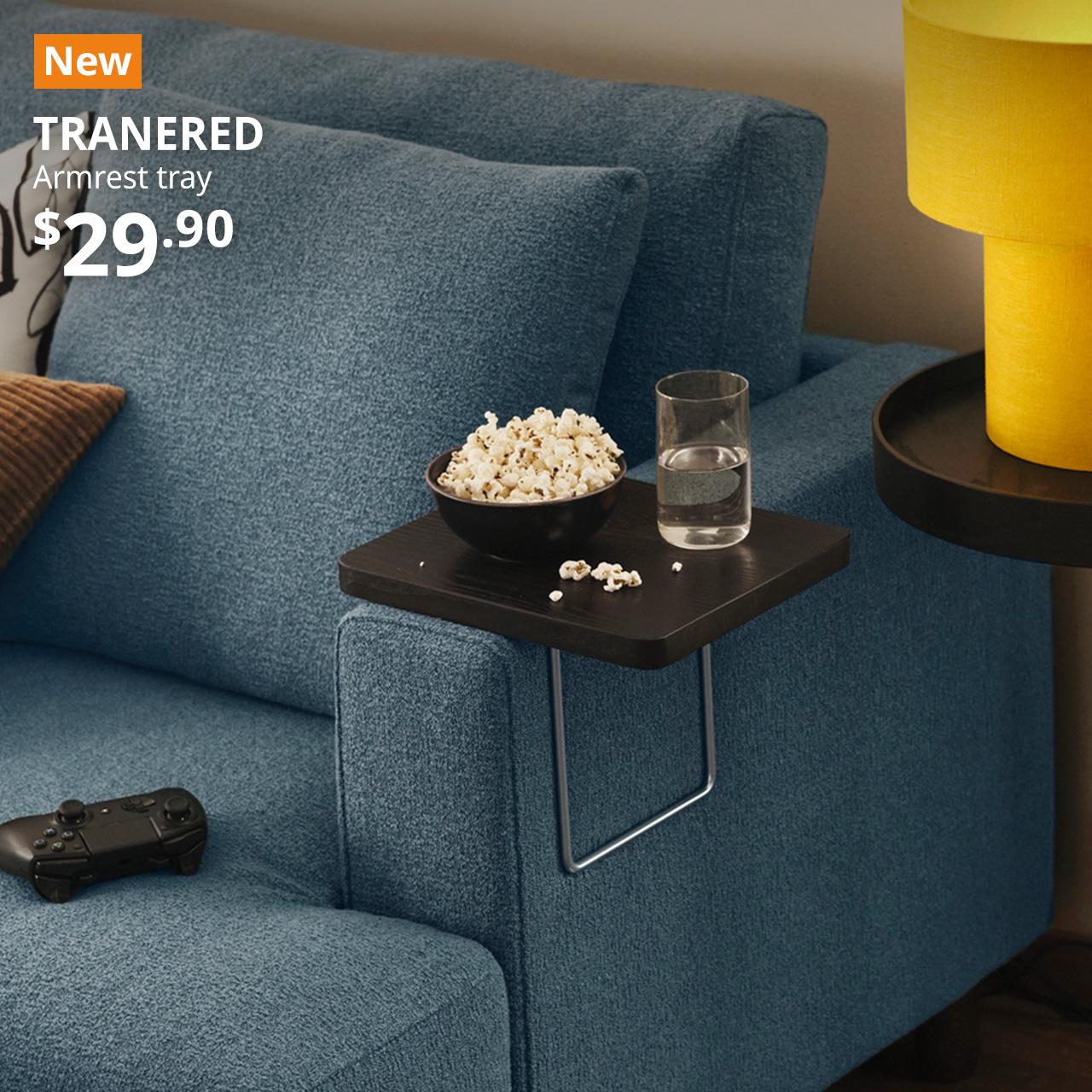 A bowl of popcorn and a glass of water placed on a dark brown TRANERED armrest tray which is clamp on a dark grey-blue UPPAKRA sofa arm in a living room seating. An orange "NEW" label is on the top left of the image with white text that reads, "TRANERED, Armrest tray, $29.90".