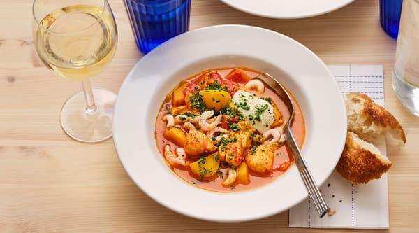 A bowl of spicy salmon stew sitting on a table next to a bun and a glass of white wine.