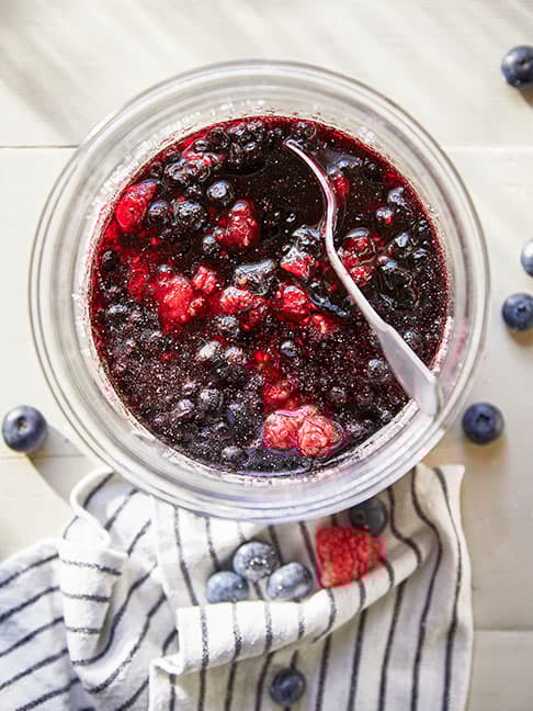 a bowl with a mixture of raspberries and blueberries in a liquid or sauce, with a spoon in it, placed on a striped cloth with some loose blueberries around it.