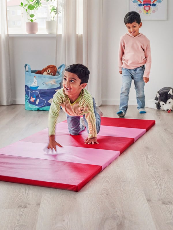A boy crawls on a pink PLUFSIG gym mat - which is unfolded and placed on the floor. His brother looks from behind and is prepared to play on the mat too. 