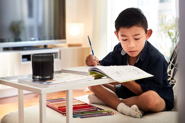 A boy is sitting on a sofa with his legs crossed, drawing in a book on a small table.