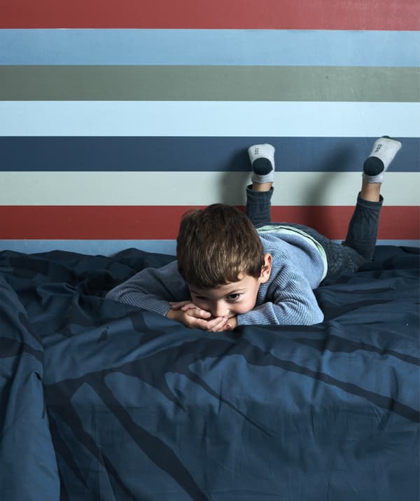 A boy lying on a bed with dark blue bedding and striped wallpaper behind.