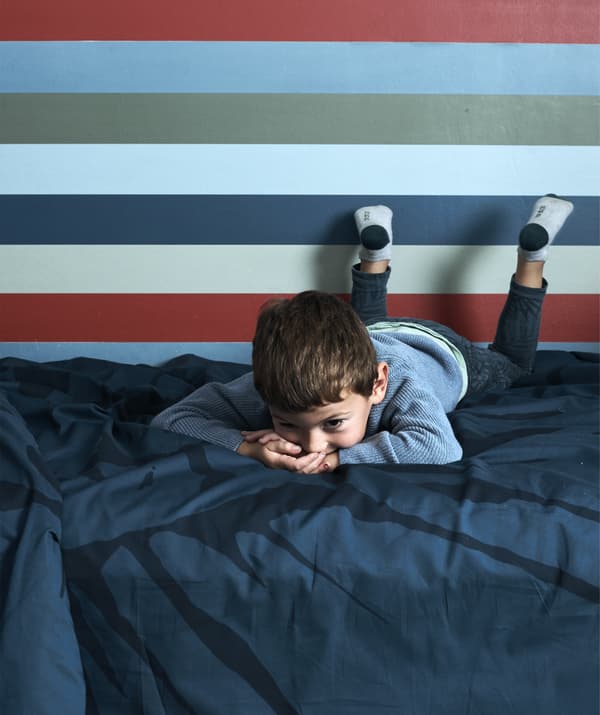 A boy lying on a bed with dark blue bedding and striped wallpaper behind.