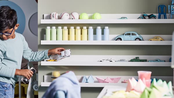 A boy organizing balls, bottles, toys, paper projects and toy cars on a wall with four white MOSSLANDA picture ledges.