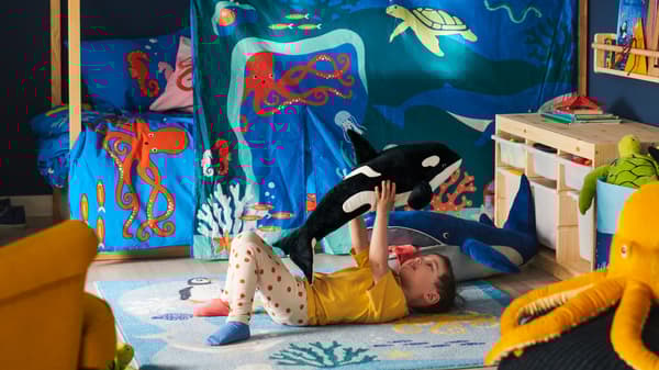 A boy plays with a BLÅVINGAD orca soft toy on a BLÅVINGAD ocean animal rug on the floor beside a KURA bed with a bed tent.