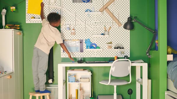 A boy standing on a stool tends to a SKÅDIS pegboard combination hung over a white PÅHL desk in a green and blue room.