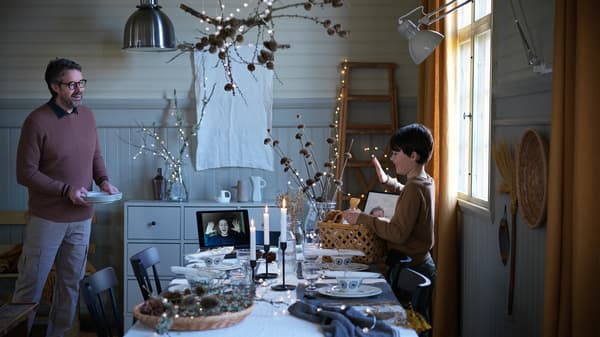 A boy waves to a person on a video call on a tablet in a candlelit festive dining room; the boy has a basket of gifts.