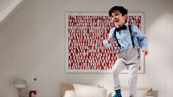 A boy wearing suspenders and a bowtie is jumping on a bed in a white room.