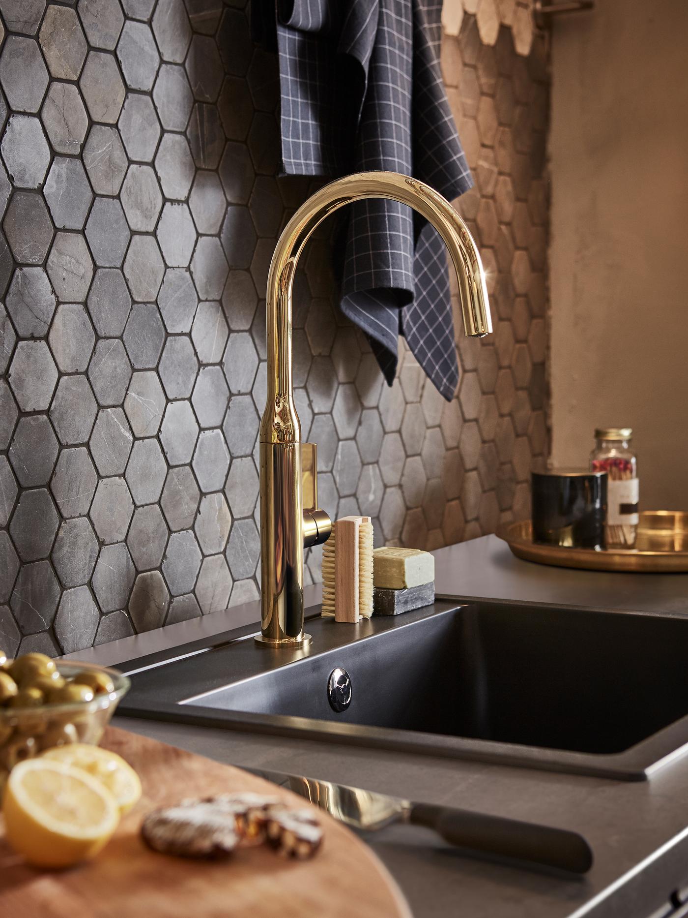 A brass-coloured mixer tap and a black quartz composite kitchen sink in front of a wall with hexagonal tiles.