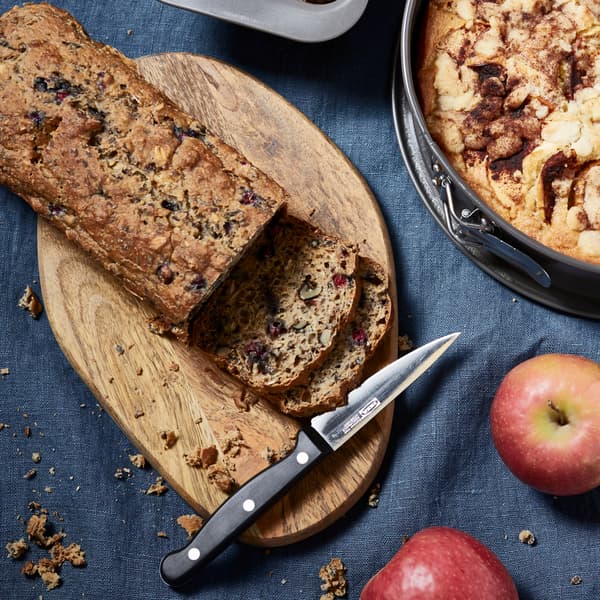 A bread that is being cut on a chopping board with a knife and some red apples.