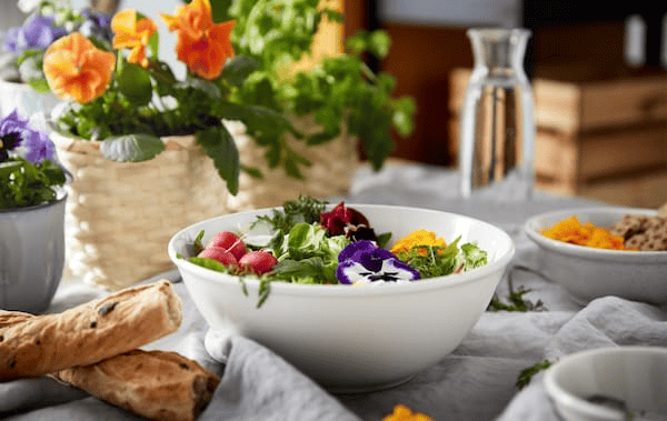 A breakfast table set with different kinds of bread, pot of flowers and a bowl of colourful garnished greens.