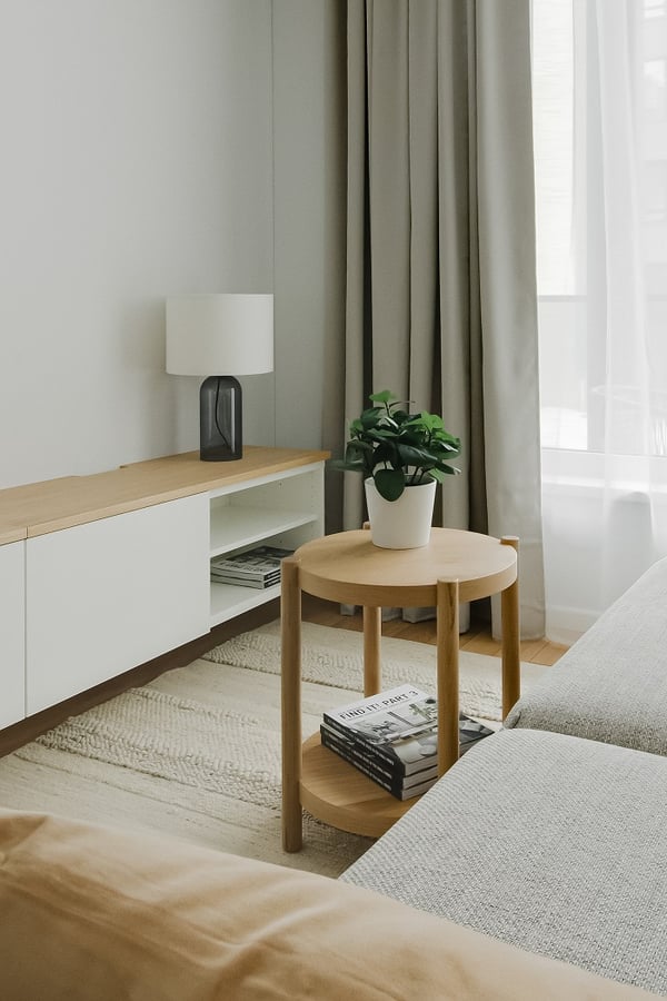 A bright and cozy living room corner with a light wooden side table holding a small green potted plant and several stacked books. Next to it stands a minimalist white and wood media console with a modern table lamp on top. Floor-to-ceiling beige curtains and sheer white drapes frame a large window, allowing soft natural light to fill the space. A textured cream rug adds warmth and comfort to the light wooden flooring.