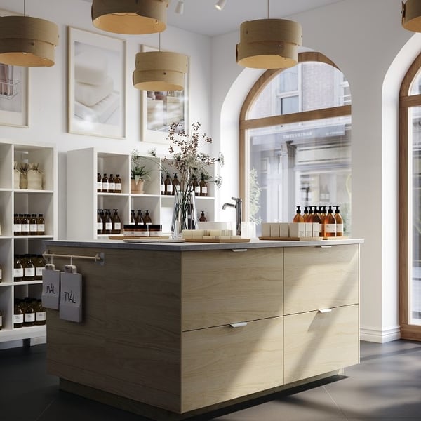 A bright and modern apothecary or skincare boutique interior. The space features a large central island with light wood drawers and a gray stone countertop, displaying amber glass bottles, soaps, and floral arrangements. Surrounding the island are white shelving units neatly stocked with brown bottles and decor items. The boutique has tall arched windows framed in wood, allowing natural light to flood the room. Above the island hang several cylindrical wooden pendant lights. Framed images of soap bars decorate the white walls, contributing to the clean and minimalist aesthetic of the store.